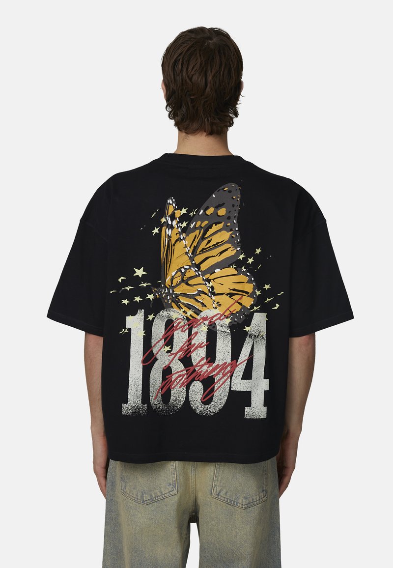 Personne portant un t-shirt noir oversize avec un grand papillon orange, des étoiles, "1984" et un texte rouge en cursive au dos, assorti à un jean clair.