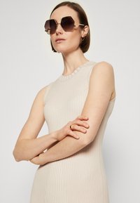 Robe sans manches beige à côtes avec une silhouette ajustée. Lunettes de soleil octogonales noires avec des accents dorés et une bague sur la main droite du mannequin.