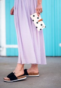 Black slide sandals with a bow detail and cork platform, paired with a lavender long dress. A white wallet with black polka dots is held in hand.