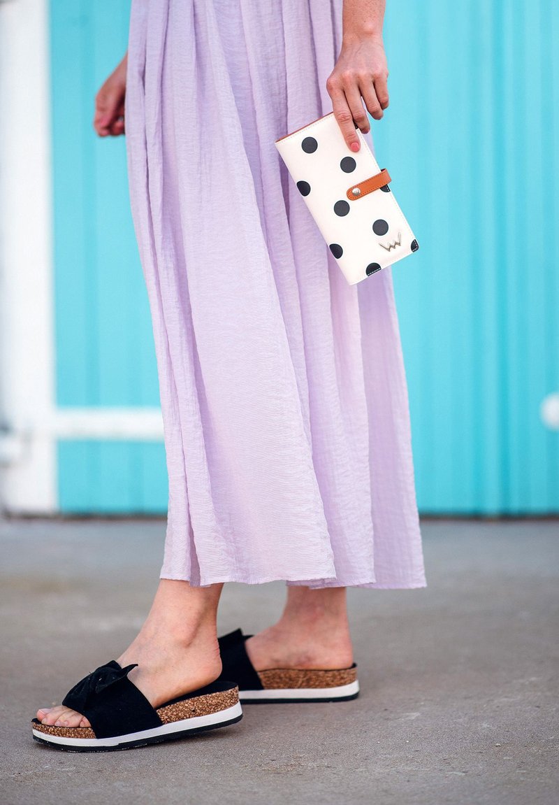 Black slide sandals with a bow detail and cork platform, paired with a lavender long dress. A white wallet with black polka dots is held in hand.