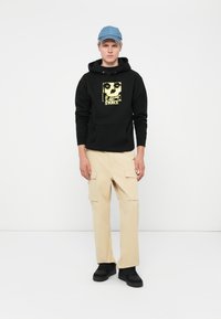 Black hoodie with a yellow skull graphic, beige cargo pants with pockets, and a light blue cap. Footwear is black. Simple, utilitarian design.