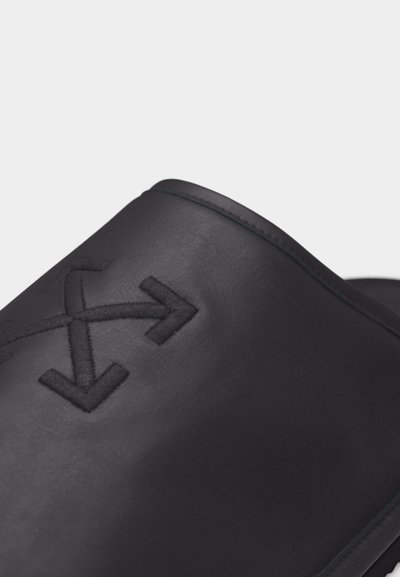 Κοντινό πλάνο μαύρου δερμάτινου σανδαλιού τύπου slide με κεντημένο λογότυπο διασταυρούμενων βελών στο πάνω λουράκι.