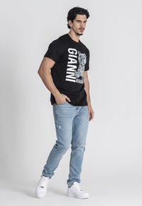 Čierne tričko s bielym textom "GIANNI KAVANI" a grafikou medveďa. Skombinované s svetlomodrými roztrhnutými džínsami a bielymi teniskami, v stoji.