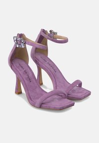 Sandalias de tacón alto de ante morado con puntera cuadrada, tiras finas y un adorno de joya en la correa del tobillo. Diseño de tacón cuadrado.