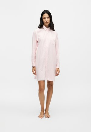 Femme aux longs cheveux foncés portant une robe-chemise rose clair à manches longues, debout pieds nus sur un fond clair uni.
