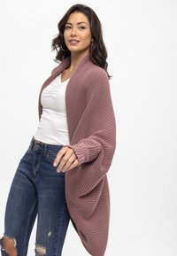 Gestrickte Strickjacke in staubigem Rose, mit einem drapierten Design, gerippter Textur und offenem Frontbereich. Getragen über einem weißen, figurbetonten Oberteil und zerrissenen Jeans.
