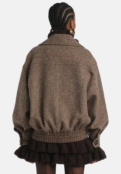 Brown herringbone jacket with a relaxed fit, oversized collar, elasticized hem, and button accents; worn over a dark ruffled skirt.