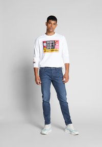 Cămașă albă cu mâneci lungi, decorată cu un imprimeu grafic multicolor, asociată cu blugi albaștri, slim-fit, și adidași albi cu accente bleumarin.