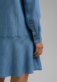 Main d'une femme reposant à côté d'une robe en jean bleu clair à manches longues avec poignets boutonnés et jupe évasée sur fond gris.