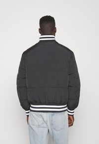Chaqueta acolchada negra con cuello acanalado, presenta rayas blancas y azul marino. Textura suave, diseño acolchado y dobladillo elástico. Vista posterior.