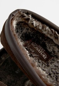 Chausson en suède marron avec une doublure douce en fausse fourrure. L'intérieur comporte une étiquette avec le texte "SIGNATURE". Forme arrondie et bords rembourrés.