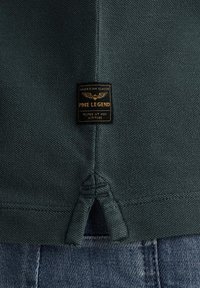 Camicia verde scuro con texture e orlo spaccato, dotata di un'etichetta nera con testo dorato che riporta "PME LEGEND" e grafica a tema aviatica.