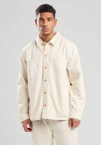 Lichtgekleurde knoopjasje gemaakt van een zachte stof. Bevat twee voorkruizen en houten knopen; losse pasvorm met een kraaguitloop.