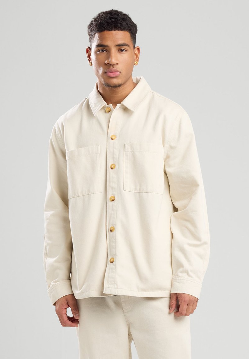 Lichtgekleurde knoopjasje gemaakt van een zachte stof. Bevat twee voorkruizen en houten knopen; losse pasvorm met een kraaguitloop.