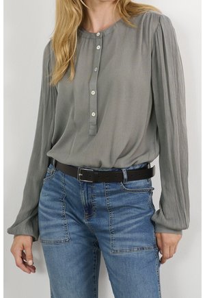 Woman wearing a loose gray blouse with long puffed sleeves and white buttons, tucked into blue jeans with large front pockets and a black belt.