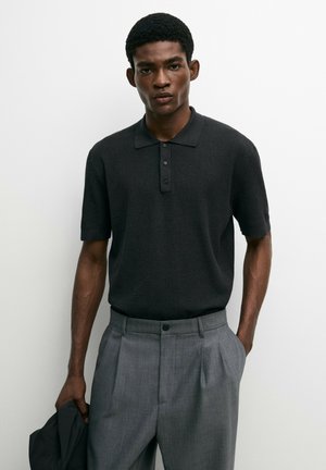 Ein Mann trägt ein schwarzes Strickpolo mit kurzen Ärmeln und graue plissierte Hosen und hält eine schwarze Jacke in der linken Hand.