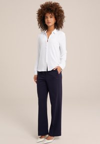 Vrouw met krullend haar, gekleed in een witte blouse met knopen, een marineblauwe broek met wijdere pijpen en witte puntige schoenen, staand met één hand in de broekzak.