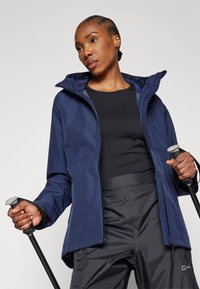 Navy waterproof jacket with a hood, side pockets, and adjustable cuffs. Worn over a black top and paired with dark hiking pants and poles.