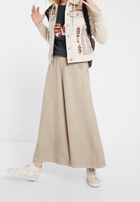 Persona que lleva pantalones beige de pierna ancha, zapatillas altas de patrón floral, una chaqueta crema con flores bordadas y una mochila negra.