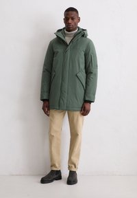 Man in een groene winterjas met capuchon, beige broek, grijze coltrui en donkere enkellaarsjes die tegen een witte muur staat.
