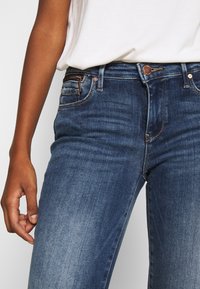 Primer plano de una persona que lleva vaqueros azules de lavado medio y una camisa blanca metida por dentro, mostrando la cintura y la parte alta del muslo.