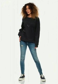 Kvinna med lockigt hår som bär en svart sweatshirt, slitna blåjeans och svarta platåskor, stående med ett ben något framåt.