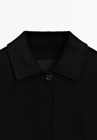 Sort uldfrakkekrave med en enkelt sort knap fastgjort øverst i midten mod en ensfarvet hvid baggrund.