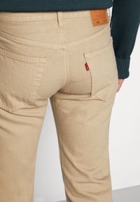 Beige denim pants featuring a five-pocket design, stitched back pockets, and a tan leather waistband label with red logo detail.