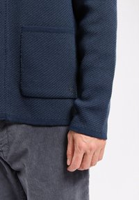 Homme portant un cardigan en tricot texturé bleu foncé avec une grande poche avant et un pantalon en velours côtelé gris, la main reposant sur le côté.