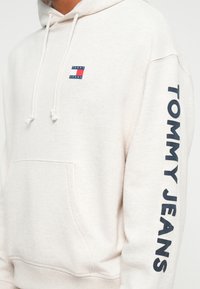 Svetlo béžová mikina s kapucňou a šnúrkami, prednou vreckom a námorníckomodrým textom "TOMMY JEANS" na rukáve. Malé logo na hrudi. Mäkko materiál.