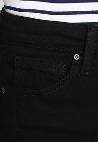 Černé džíny z denimu s přední kapsou a jemným prošitím a stříbrným kovovým nýtem. Materiál je mírně strukturovaný a odolný.