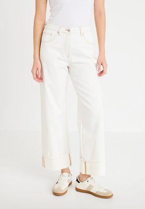 Persoon die witte jeans met wijde pijpen en hoge taille draagt, met omgeslagen zomen en beige-witte sneakers, staand tegen een effen witte achtergrond.