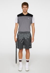 Grå atletisk polo med svarta och vita detaljer, ihop med glansiga grå shorts, vita sneakers och mönstrade strumpor, håller en tennisracket.