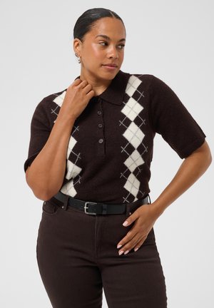 Femme portant un pull à manches courtes marron avec un motif losanges blanc et un pantalon marron foncé, regardant sur le côté avec la main près du col.