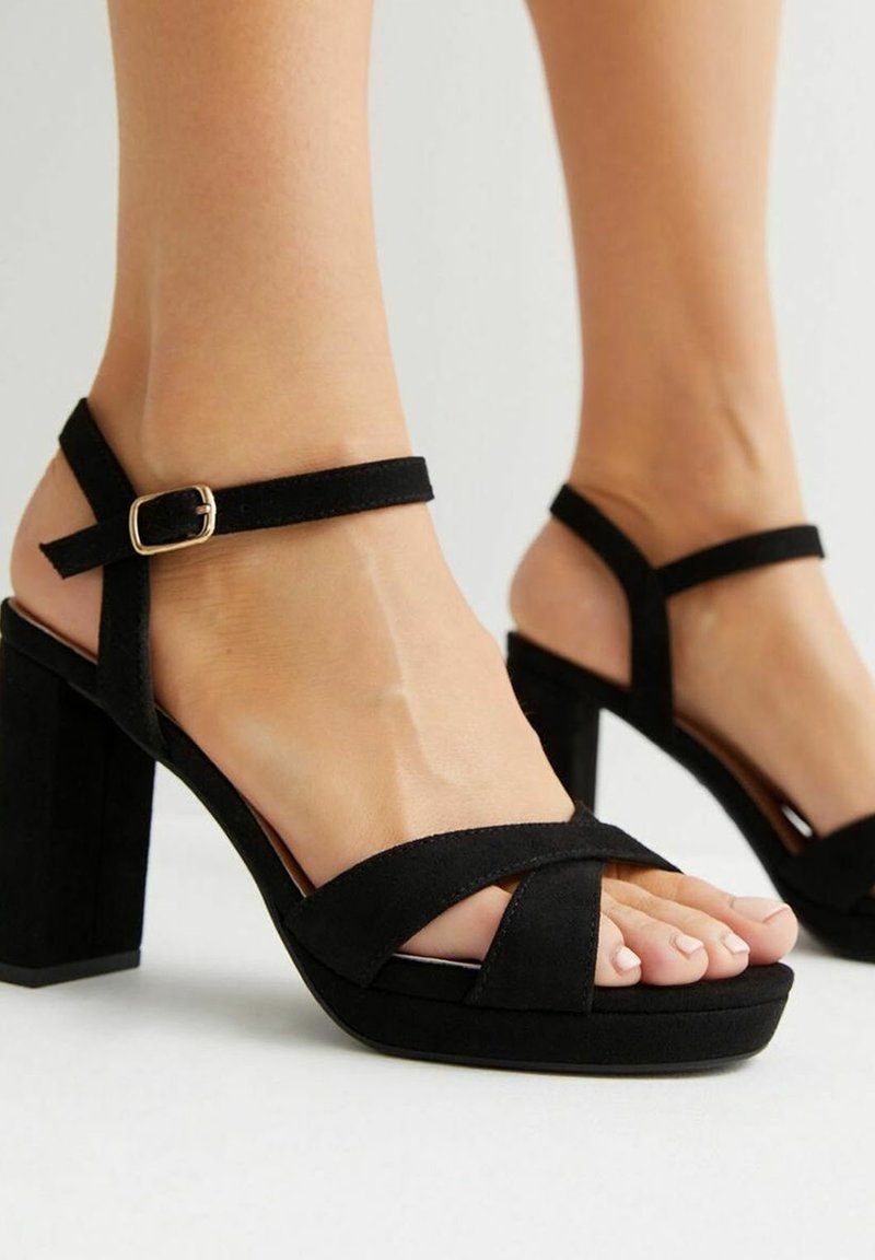 Pieds portant des sandales à talons blocs en daim noir à bout ouvert, avec brides à la cheville et fermetures à boucle dorée sur un fond clair.