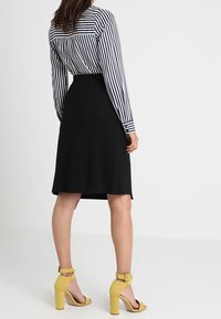 Jupe noire avec une taille ajustée et un ourlet au niveau des genoux, associée à une chemise à manches longues rayée noir et blanc et des sandales à talons hauts jaunes.