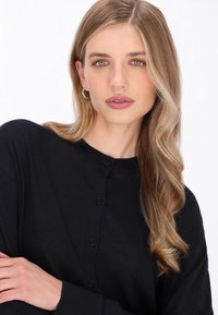 Cardigan negru tricotat cu guler rotund și închidere cu nasturi. Textură fină, cu mâneci lungi și o croială relaxată. Modelul evidențiază caracteristicile acestuia.