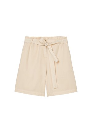 Femme portant un polo blanc à manches courtes, un short beige taille haute avec une ceinture nouée, et des chaussons blancs pelucheux à bout ouvert.