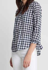 Camisa de cuadros en azul marino y blanco, con mangas remangadas, fabricada con un tejido ligero y un dobladillo curvo. Presenta bolsillos frontales y botones de madera.