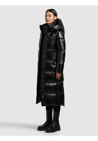 Sieviete, kas valkā garu melnu spīdīgu pītas mēteli ar kapuci, melnas legingas un masīvas melnas botas, stāvot pret vienkāršu gaišu fonu.