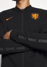 Zwarte zip-up jack gemaakt van gladde stof. Heeft een oranje Nike-logo en een wapenschild met een leeuw. "KNVB" is gedrukt langs de mouwen.