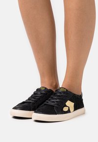 Baskets noires avec une finition texturée, dotées d'une semelle en caoutchouc crème et d'un accent beige stylisé sur le côté. Les lacets sont noirs.