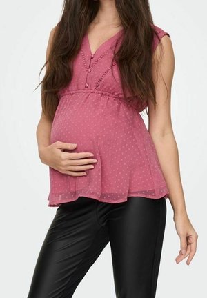 Femme enceinte portant un chemisier rose sans manches et un pantalon noir, tenant doucement son ventre d'une main, debout devant un fond blanc.