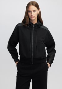 Sudadera negra cortada de tejido suave, con cierre de cremallera, capucha con cordón y paneles de malla en los hombros.