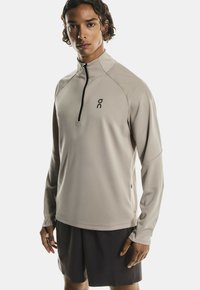 Smėlinis ilgomis rankovėmis sportinis marškinėliai su pusiau užtrauktuku priekyje, turintys juodą logotipą krūtinėje ir tekstūruotą paviršių oro pralaidumui.