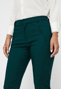 Turkusowe spodnie stretchingowe o dopasowanym kroju, z gładkimi kieszeniami z przodu i szlufkami na pasek, w połączeniu z białą koszulą z guzikami.