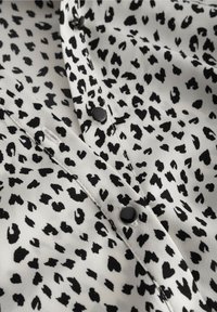 Blouse noire et blanche présentant un motif tacheté, réalisée en tissu doux. Elle comporte des boutons noirs sur le devant et des surpiqûres détaillées.