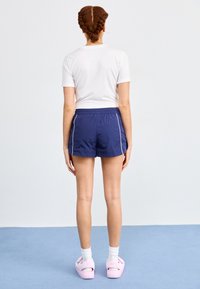 Jūriniai sportiniai šortai su elastinga juosta ir baltais šonų juostelėmis, derinami su balta prigludusia marškinėliais ir rožinėmis šlepetėmis.