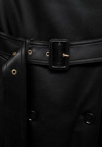 Ceinture en cuir noir avec une boucle rectangulaire attachée autour d'un vêtement en cuir noir texturé avec des boutons visibles.