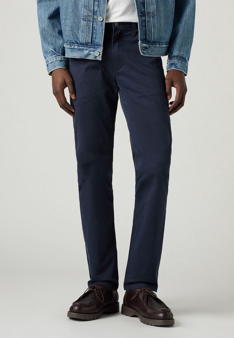 Homme portant un pantalon bleu marine, des chaussures en cuir marron foncé, une chemise blanche et une veste en denim bleue, se tenant devant un fond uni.